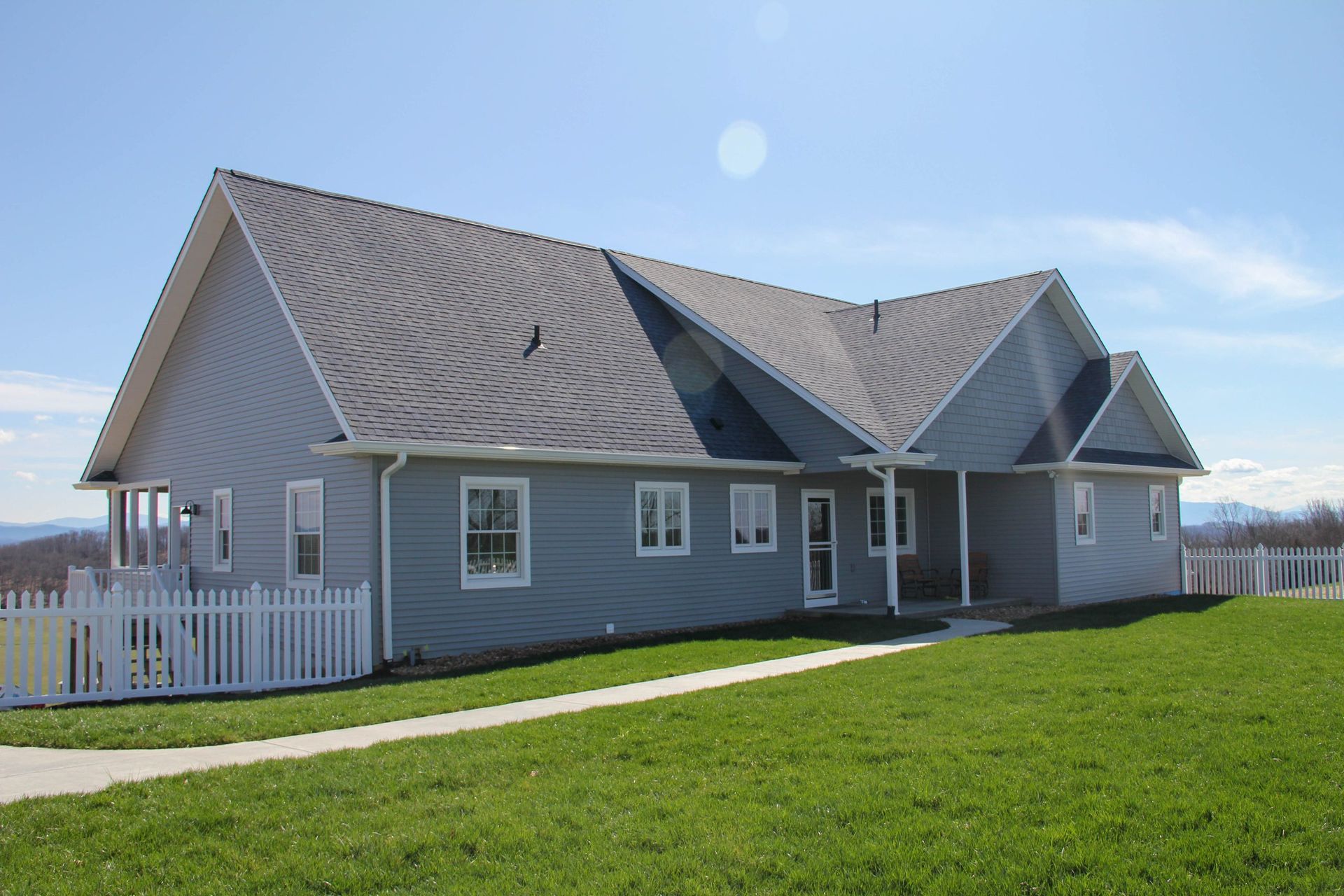 A gray, ranch-style house with a light gray shingled roof, a white picket fence, and a green lawn under a clear blue sky.