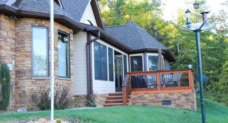 A house with stone and beige siding, a screened porch, and a wood deck, surrounded by green trees on a grassy lawn.