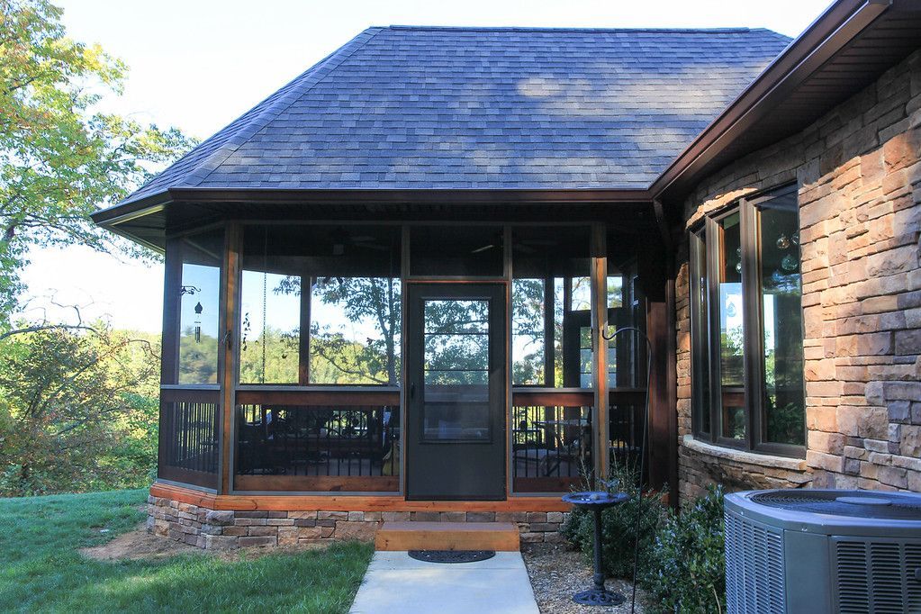 A screened-in porch with a dark wood frame attached to a brick house next to a yard with a concrete walkway.