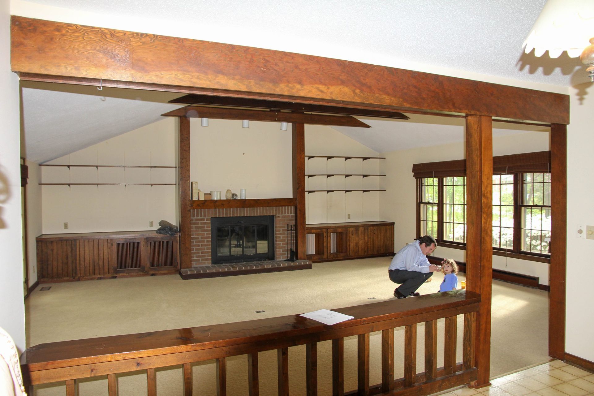 A person kneels to interact with a child in a large, carpeted living room featuring wood trim and a brick fireplace.