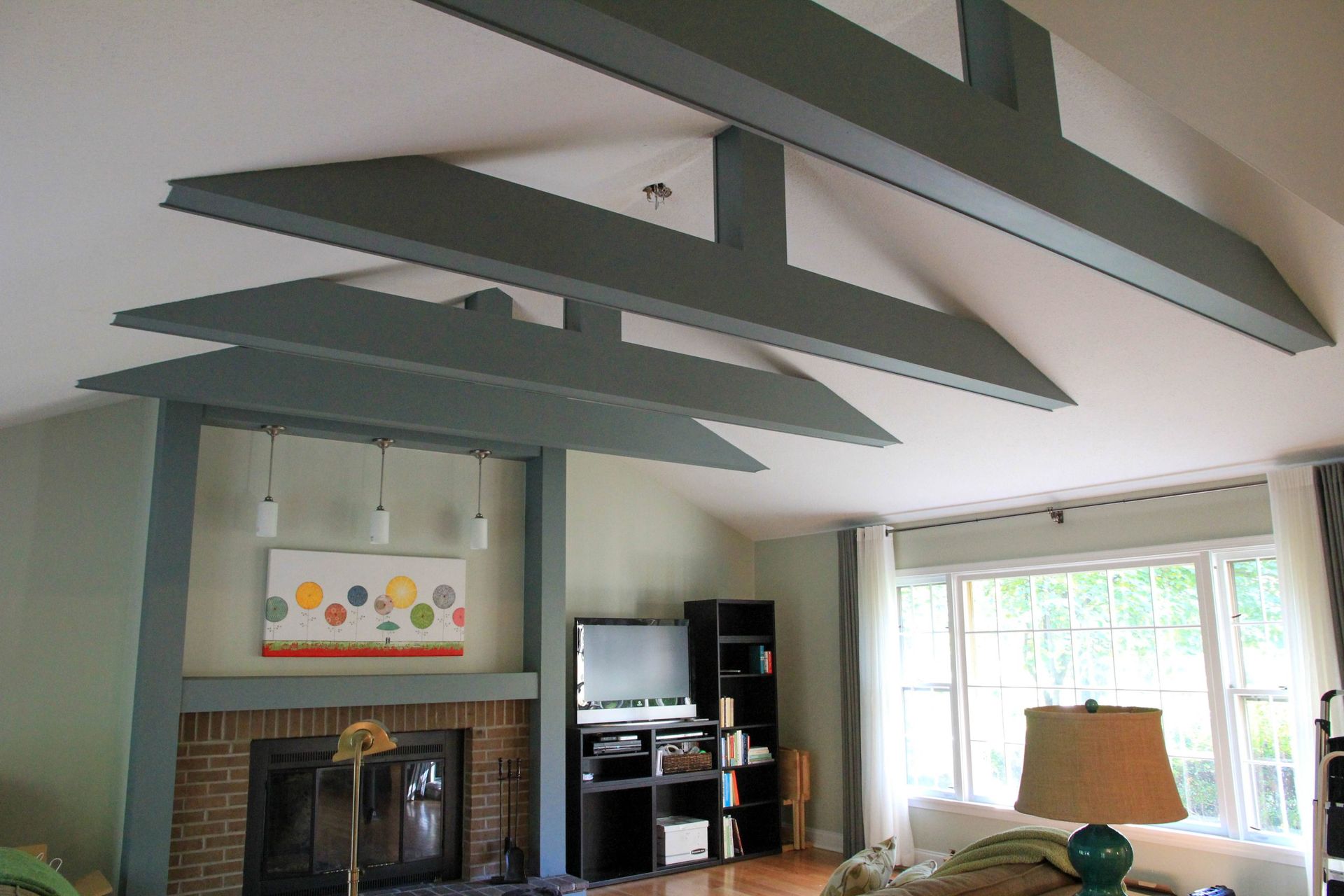 A living room features a brick fireplace, a television on a dark shelf, and three dark gray wooden ceiling beams.