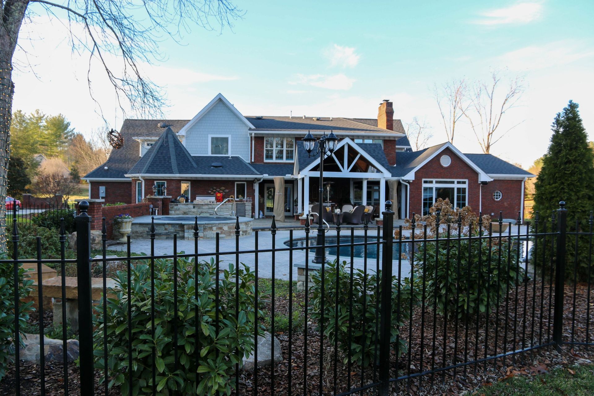 A large multi-story home with red brick and grey siding features a pool, patio, and black metal fence in the backyard.