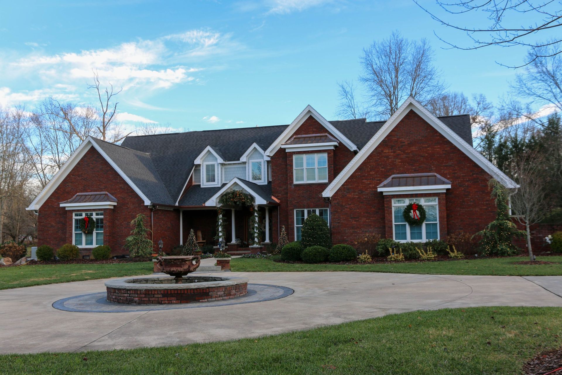 A large two-story brick house with multiple gables, a circular driveway, and a central stone fountain in the front yard.