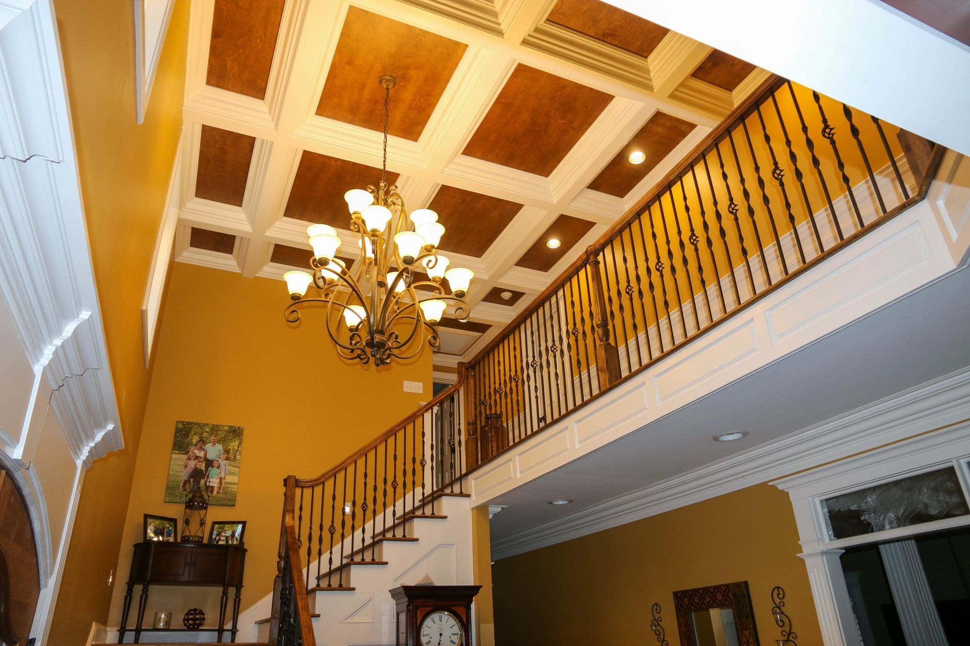 A high-ceilinged foyer with a coffered wooden ceiling, a large chandelier, a staircase with railing, and yellow walls.
