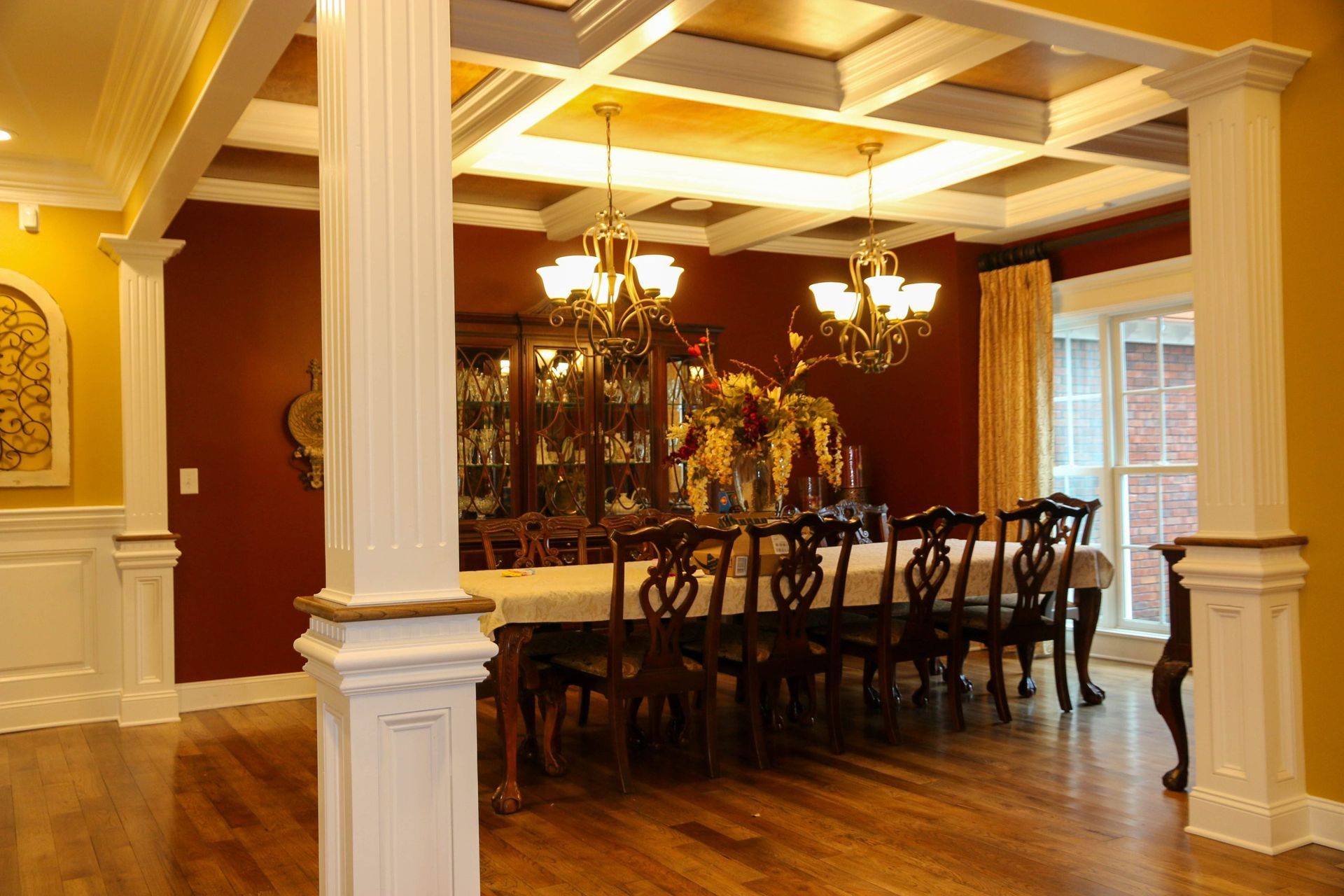 A dining room with a long table, dark red walls, two chandeliers, and a coffered ceiling between two white pillars.