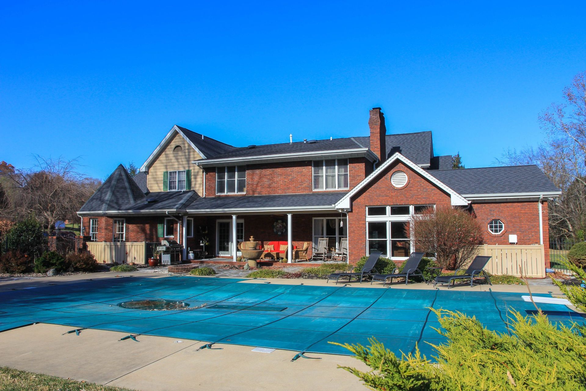 A red brick house with a dark roof and a pool covered by a blue tarp, set against a clear blue sky.