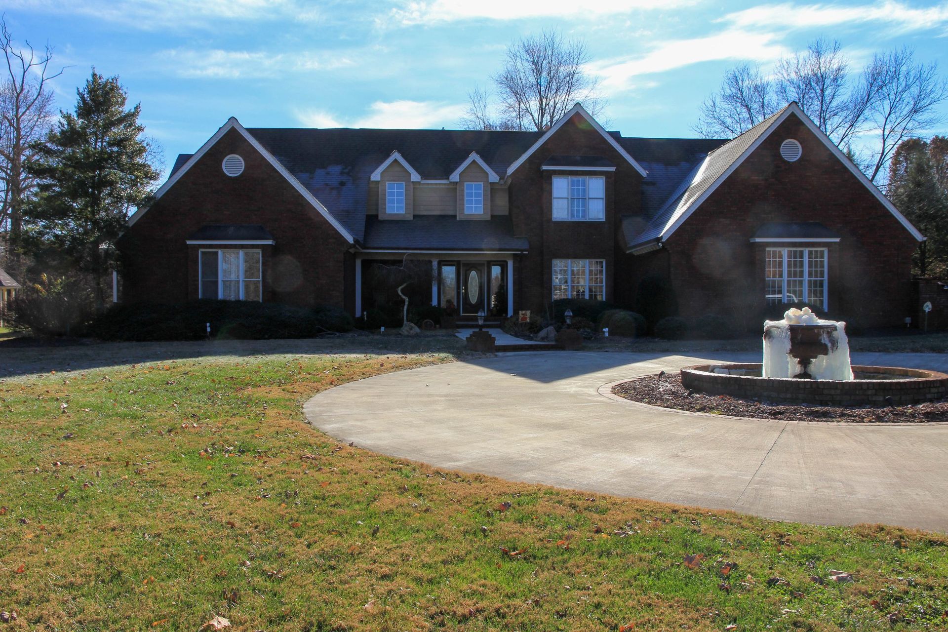Two-story brick house with a circular driveway and stone fountain in a grassy front yard under a blue sky.