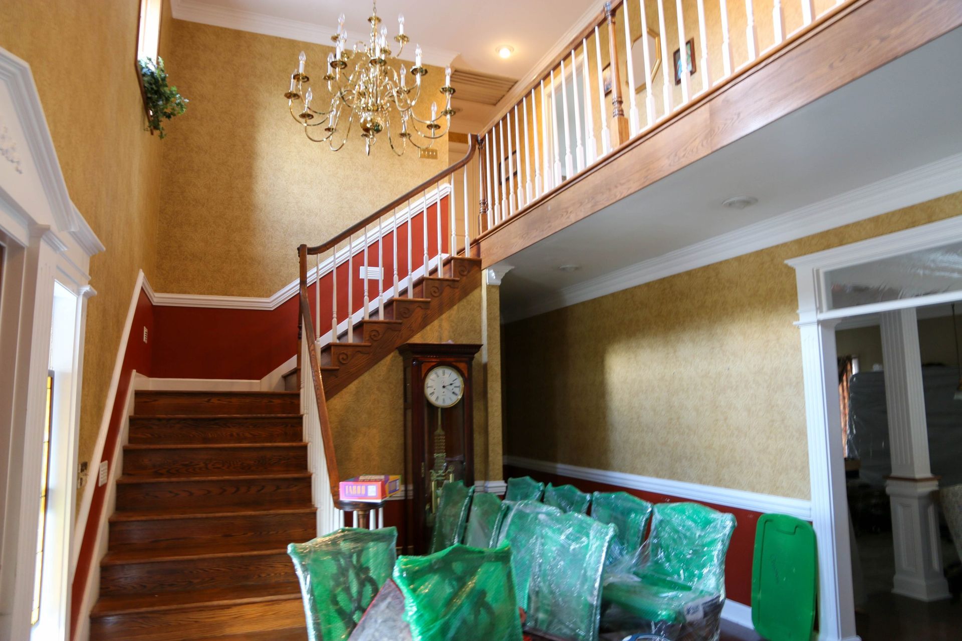 Foyer with a wooden staircase, gold patterned wallpaper, a chandelier, a grandfather clock, and chairs wrapped in plastic.