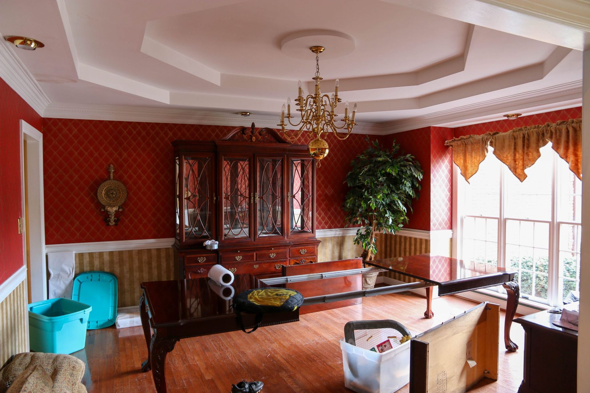 A dining room with red patterned walls, wood furniture, a chandelier, and a large window with gold valances.