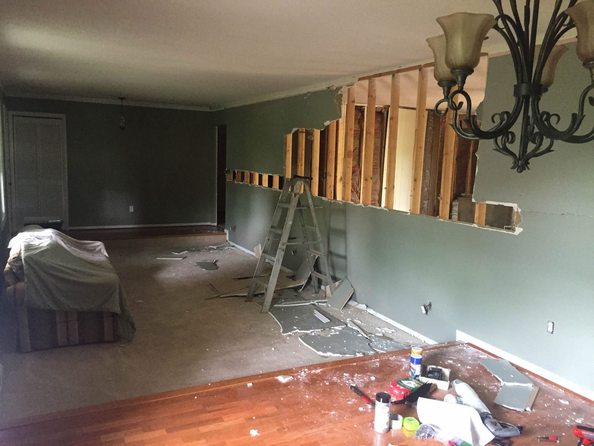 An interior room under renovation with a hole in a dark green wall, wooden studs exposed, a stepladder, and debris.