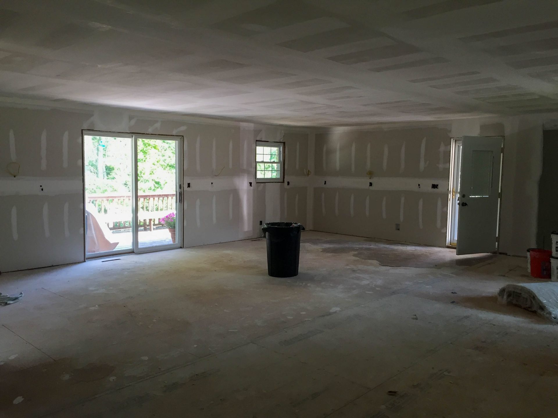 An interior room under construction with bare drywall walls and ceiling, a glass sliding door, and a black trash bin.
