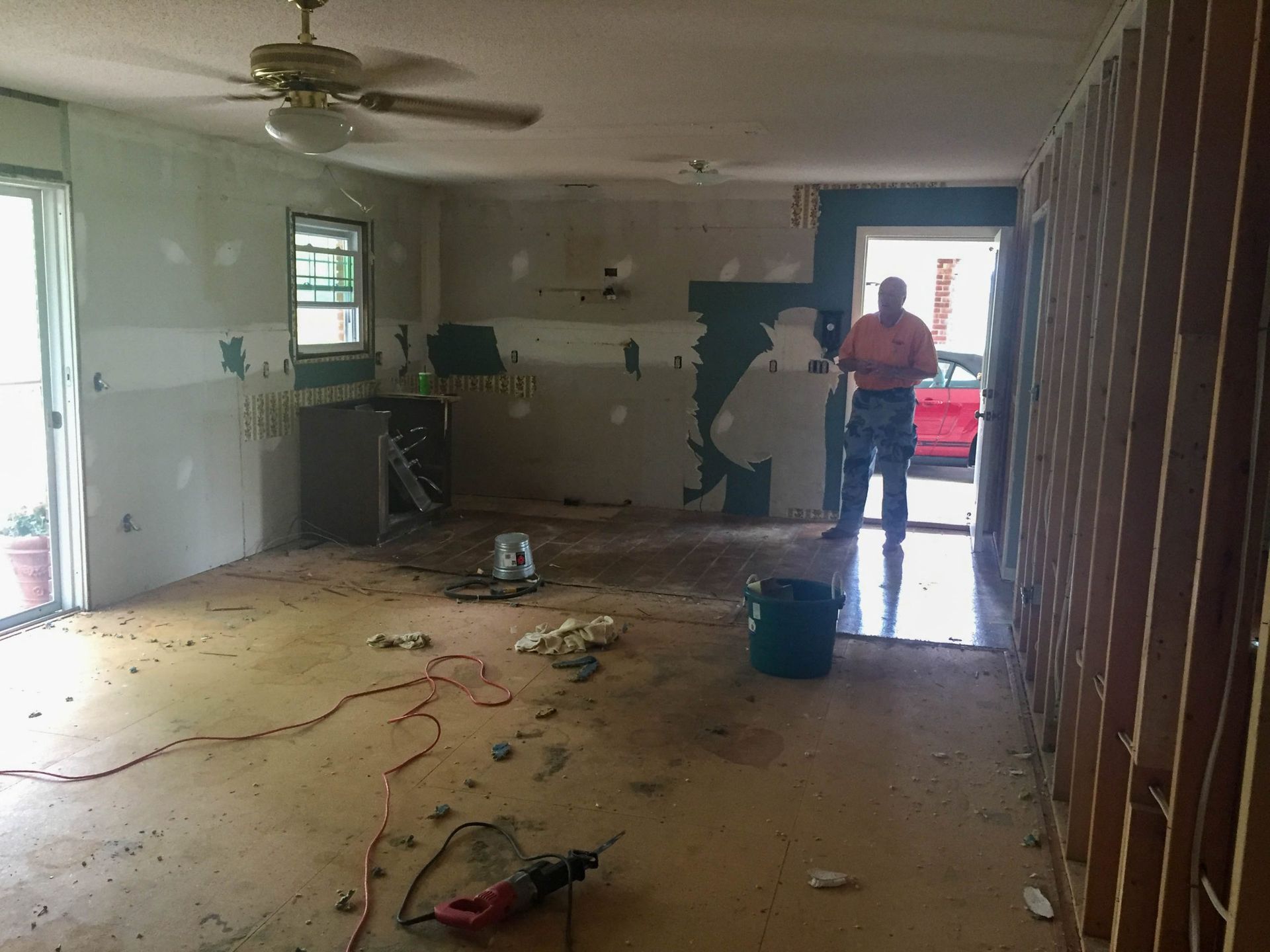 A room under construction with exposed wall studs, scattered debris, and a person standing in an open doorway.