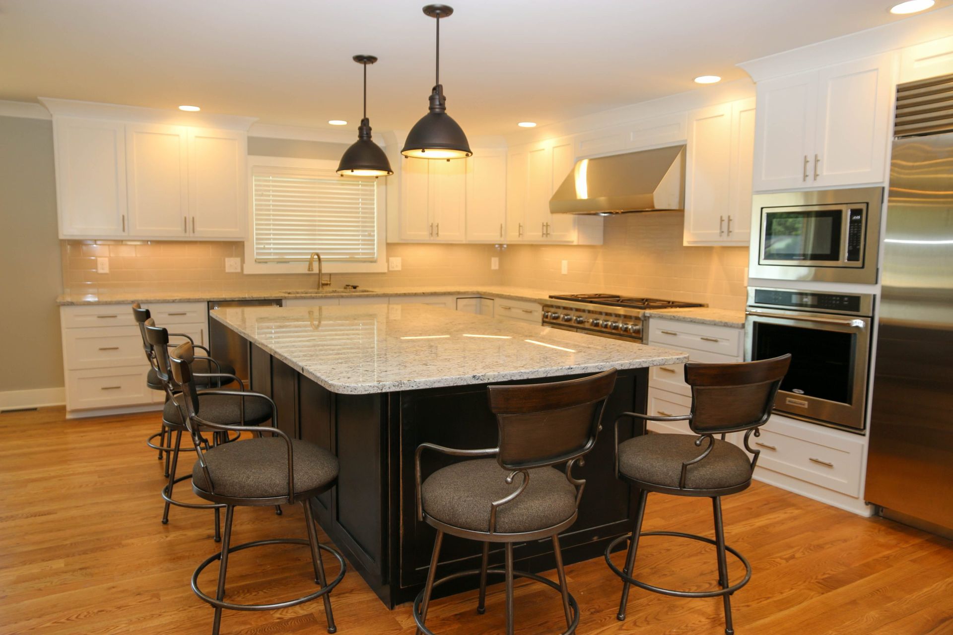 A kitchen with white cabinets, a black island, granite countertops, bar stools, stainless appliances, and pendant lights.