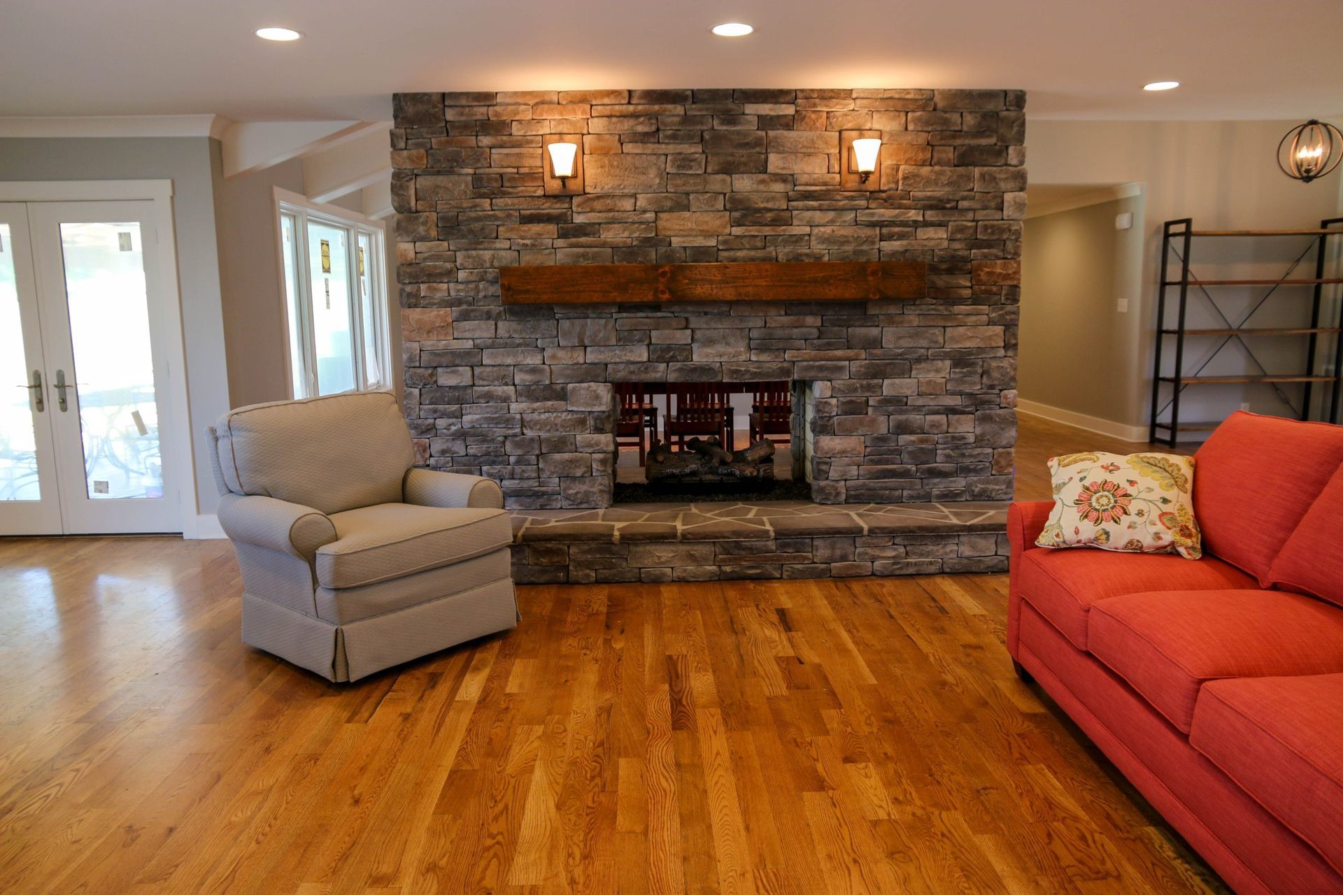 A stone fireplace with a wooden mantel in a living room featuring a light chair, a red sofa, and hardwood floors.