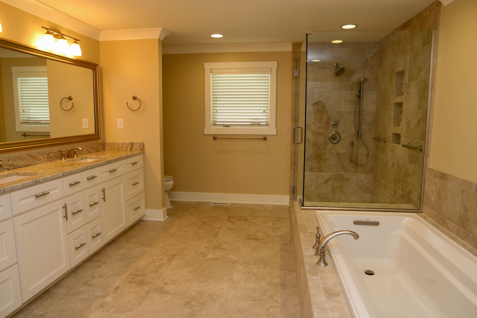 A modern bathroom with a white double vanity, beige tile floor, a glass shower, and a built-in soaking tub.