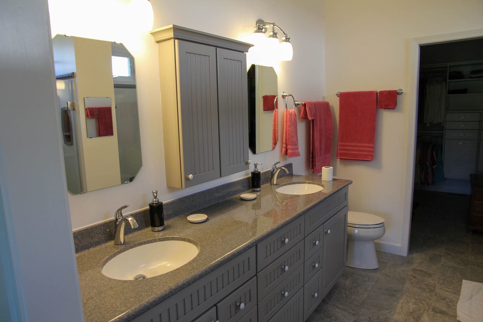 A double-sink bathroom vanity with grey cabinets, granite countertops, red towels, and a toilet in the corner.