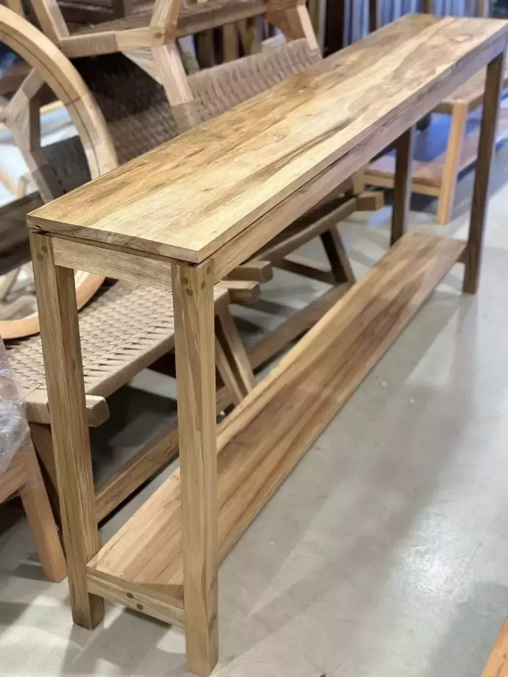 Consola de madera con estante tejido, rodeada de sillas de madera en un taller.