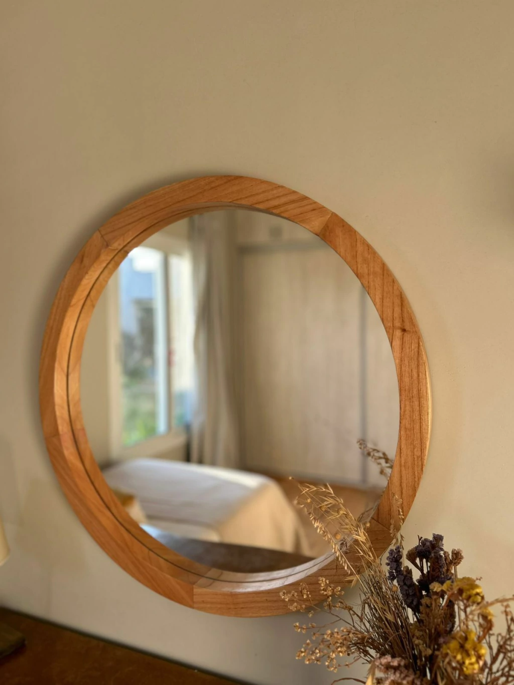 Espejo redondo con marco de madera en una pared, que refleja una cama y una ventana con una planta delante.