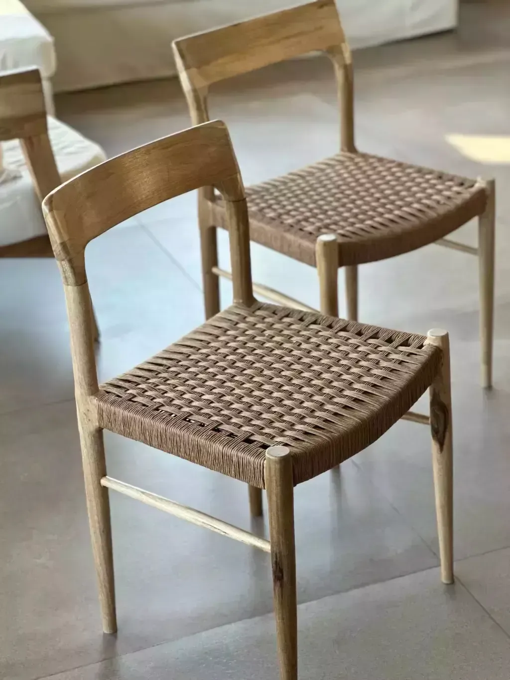 Dos sillas de madera clara con asientos tejidos, sobre un piso de baldosas.