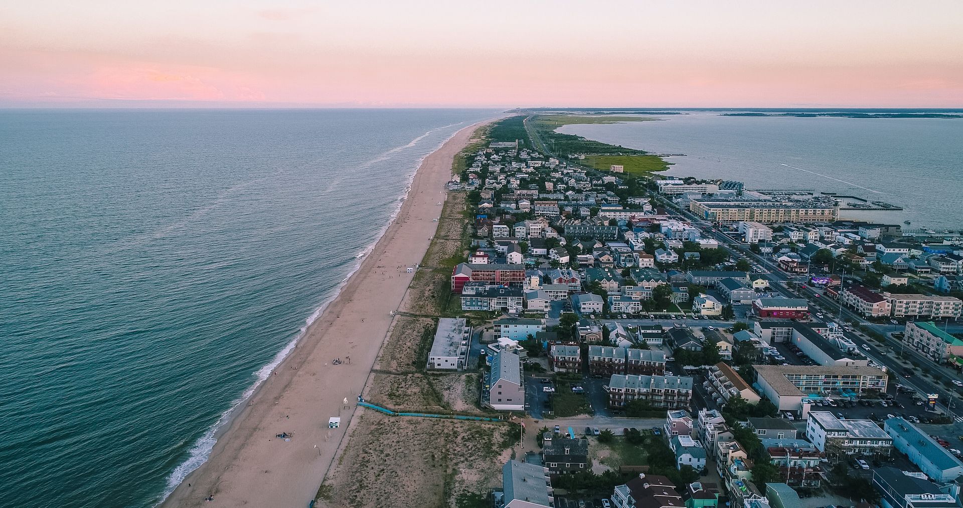 Aerial view of a coastal town and long beach at sunset, with blue ocean and pink sky.