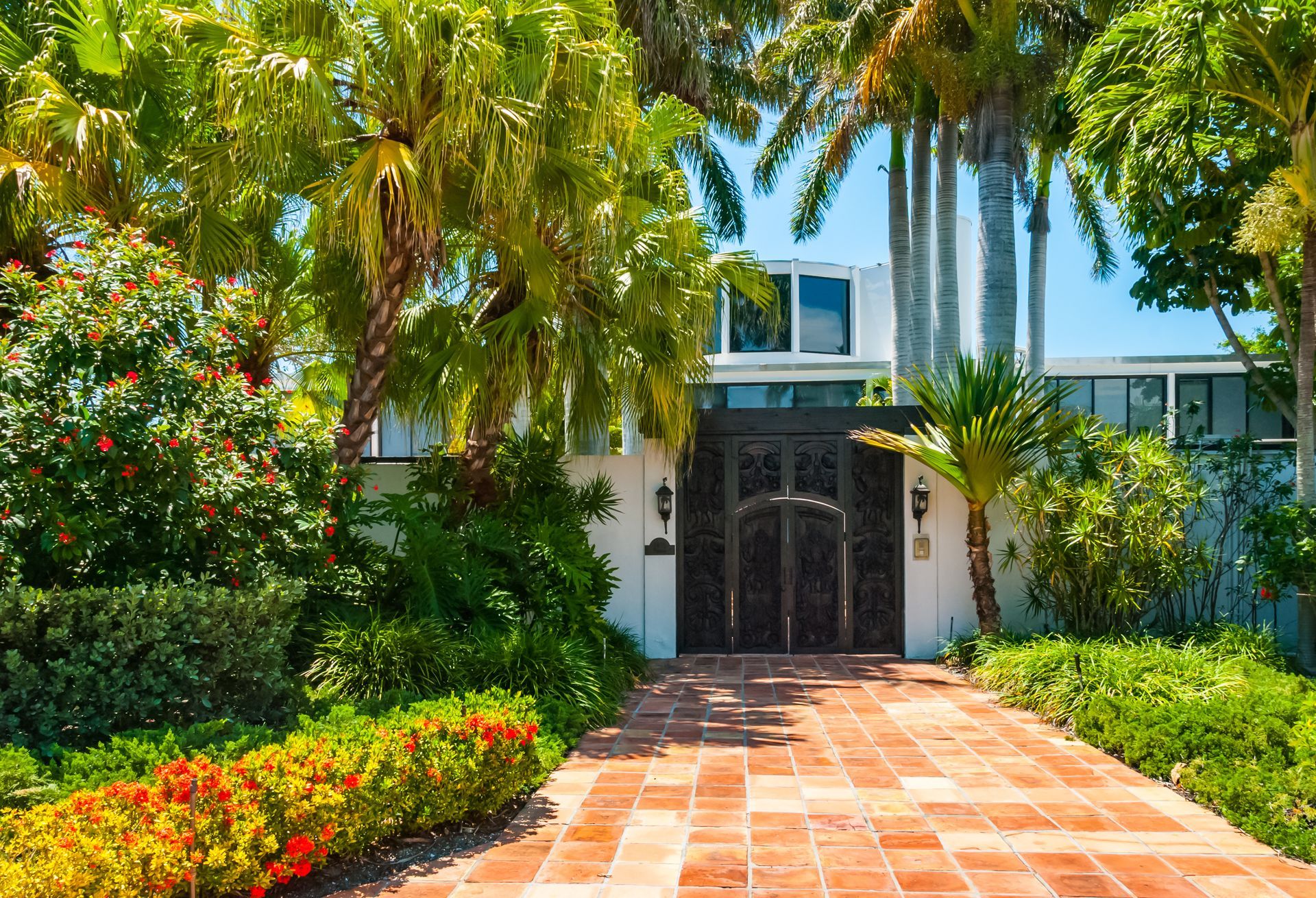 Brick path leads to an ornate black gate in front of a white building, surrounded by lush tropical landscaping.