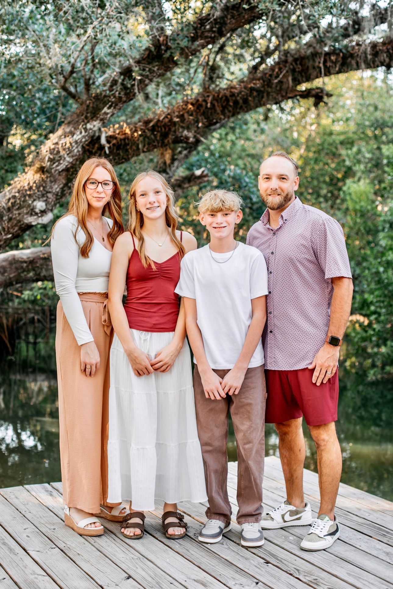 A family of four stands together on a wooden pier in front of a tree and water, smiling at the camera.
