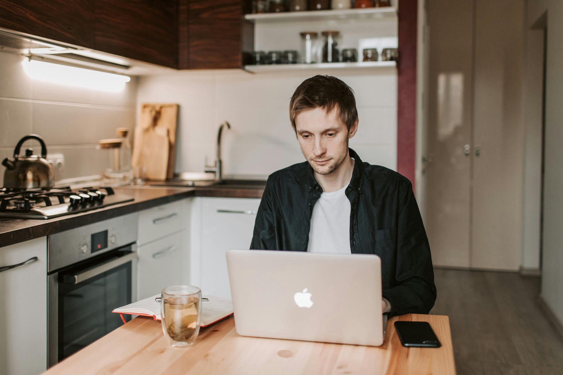 Man working on laptop at a wooden table in a kitchen. Cup of tea, phone, and notebook present.