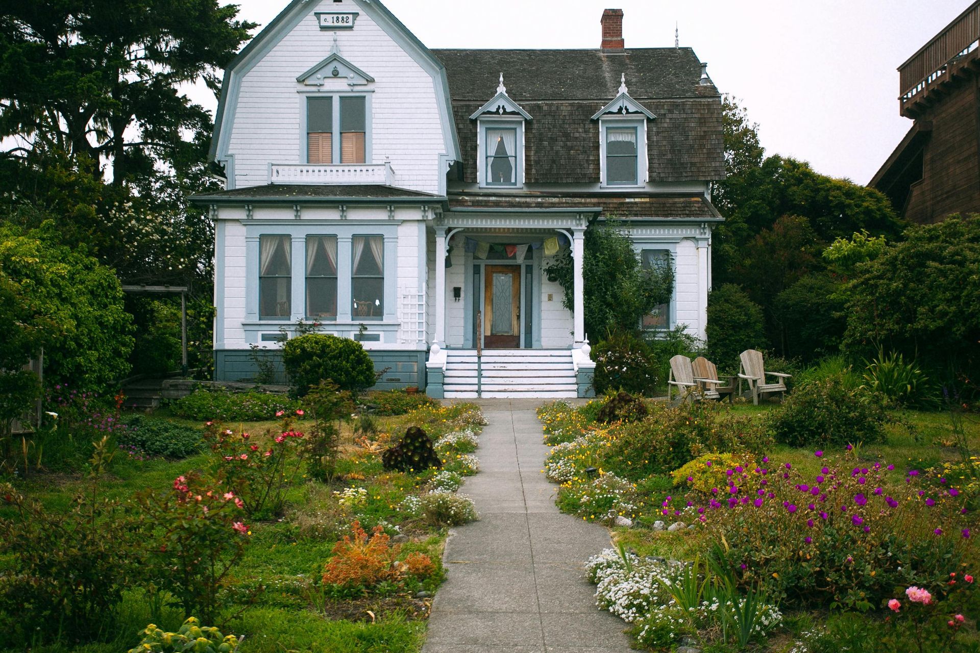 White Victorian house with blue trim, surrounded by a lush garden and a pathway leading to the front door.