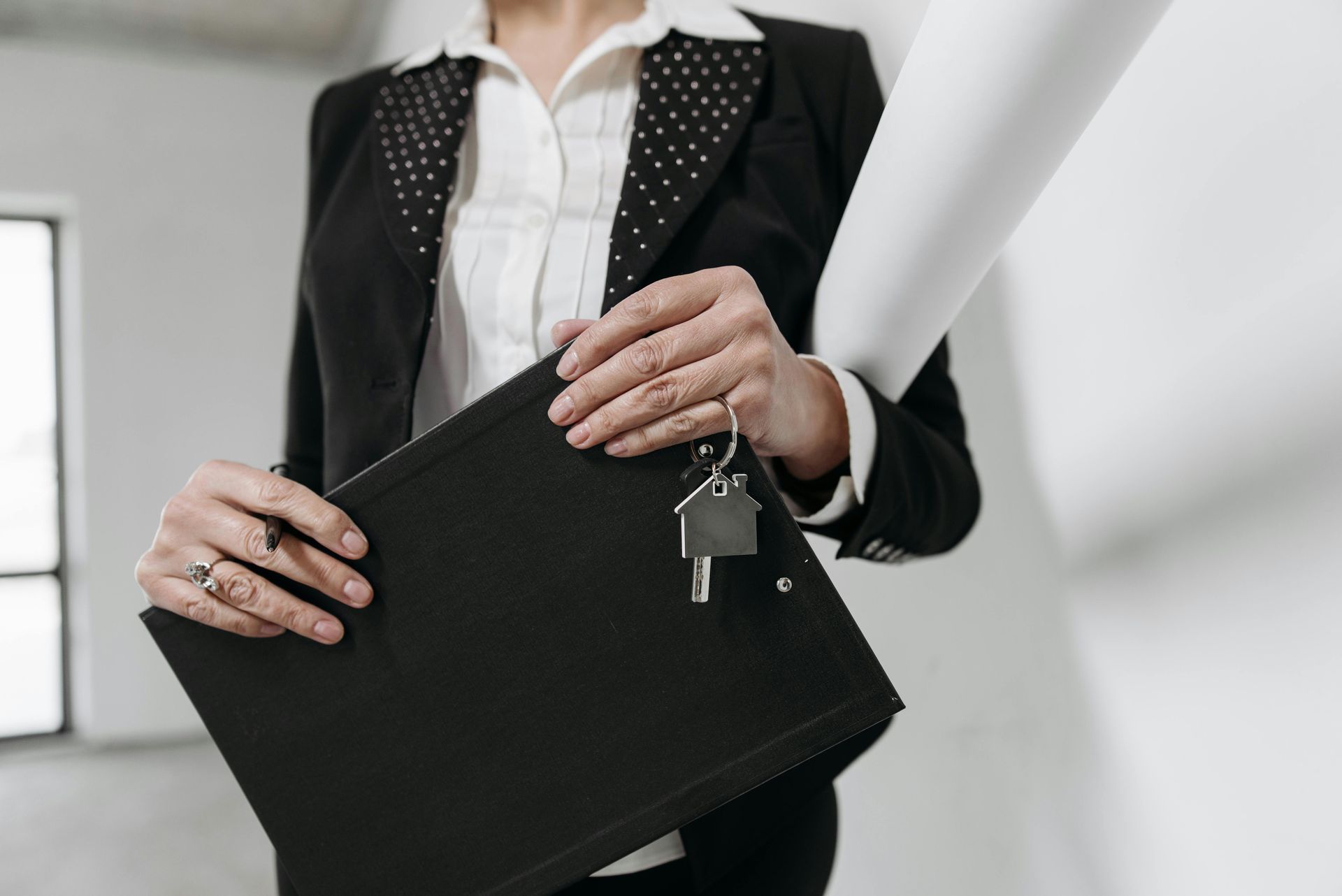 Woman in a suit holding keys with house charm, a rolled blueprint, and a black file folder.