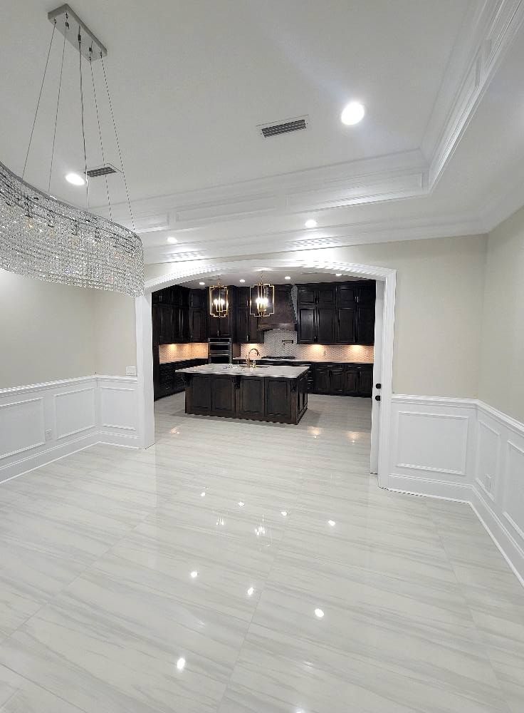 A large empty room with a kitchen in the background and a chandelier hanging from the ceiling.