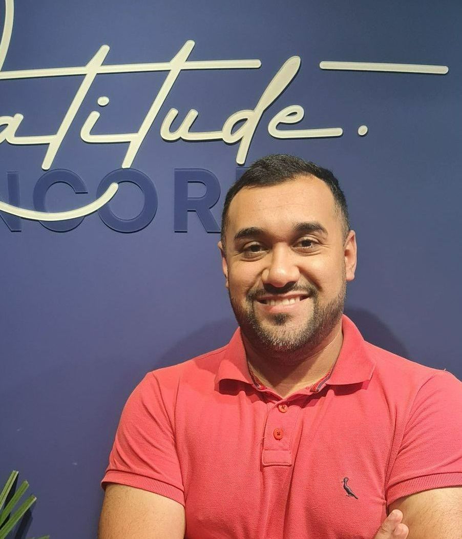 Um homem de camisa azul está sorrindo com os braços cruzados em frente a uma parede roxa.