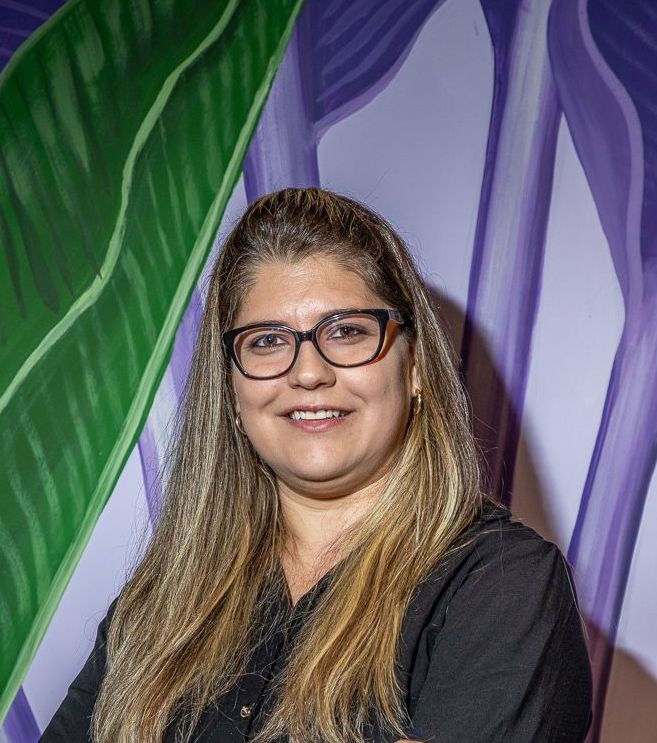 Uma mulher de óculos e camisa preta sorri diante de uma pintura de flores.