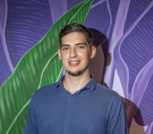 Um homem de camisa azul está parado em frente a uma parede roxa com uma folha verde.