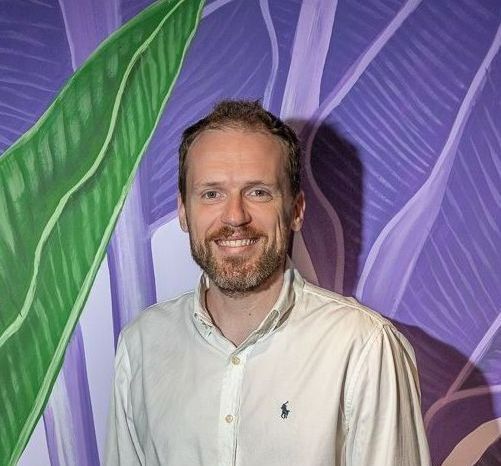 Um homem com barba está sorrindo em frente a uma parede roxa.