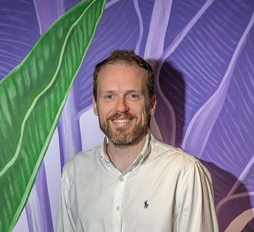 Um homem com barba está sorrindo em frente a uma parede roxa.