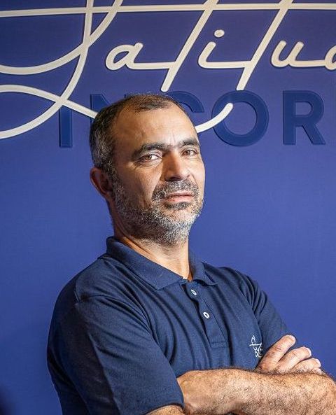 Um homem barbudo está parado com os braços cruzados em frente a uma parede azul.