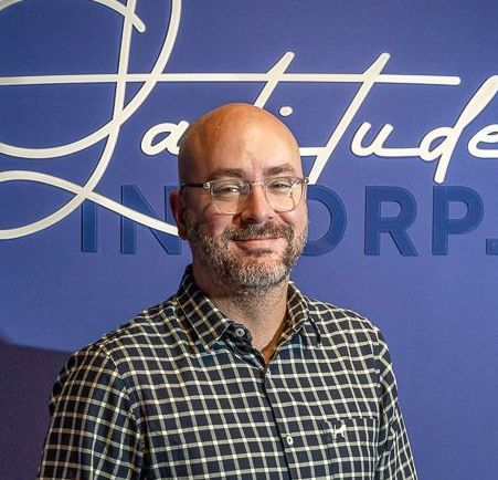 Um homem careca com óculos e barba está parado em frente a uma parede azul.