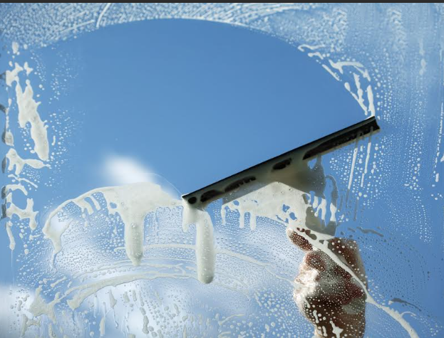 Hand using a squeegee to clean a soapy window, revealing blue sky.