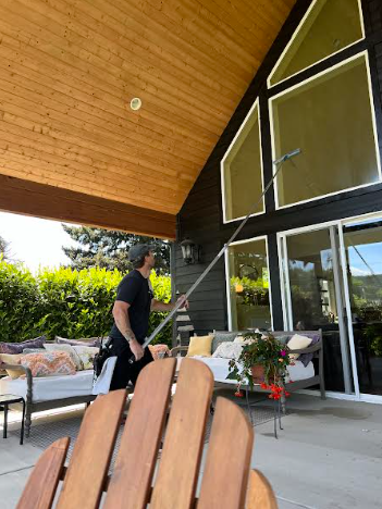 Man cleaning windows with a pole from a patio. Black house with tall windows, wooden ceiling.