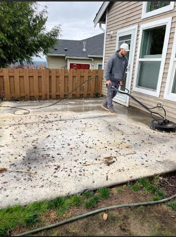 Person pressure washing a concrete patio next to a house with a wooden fence.