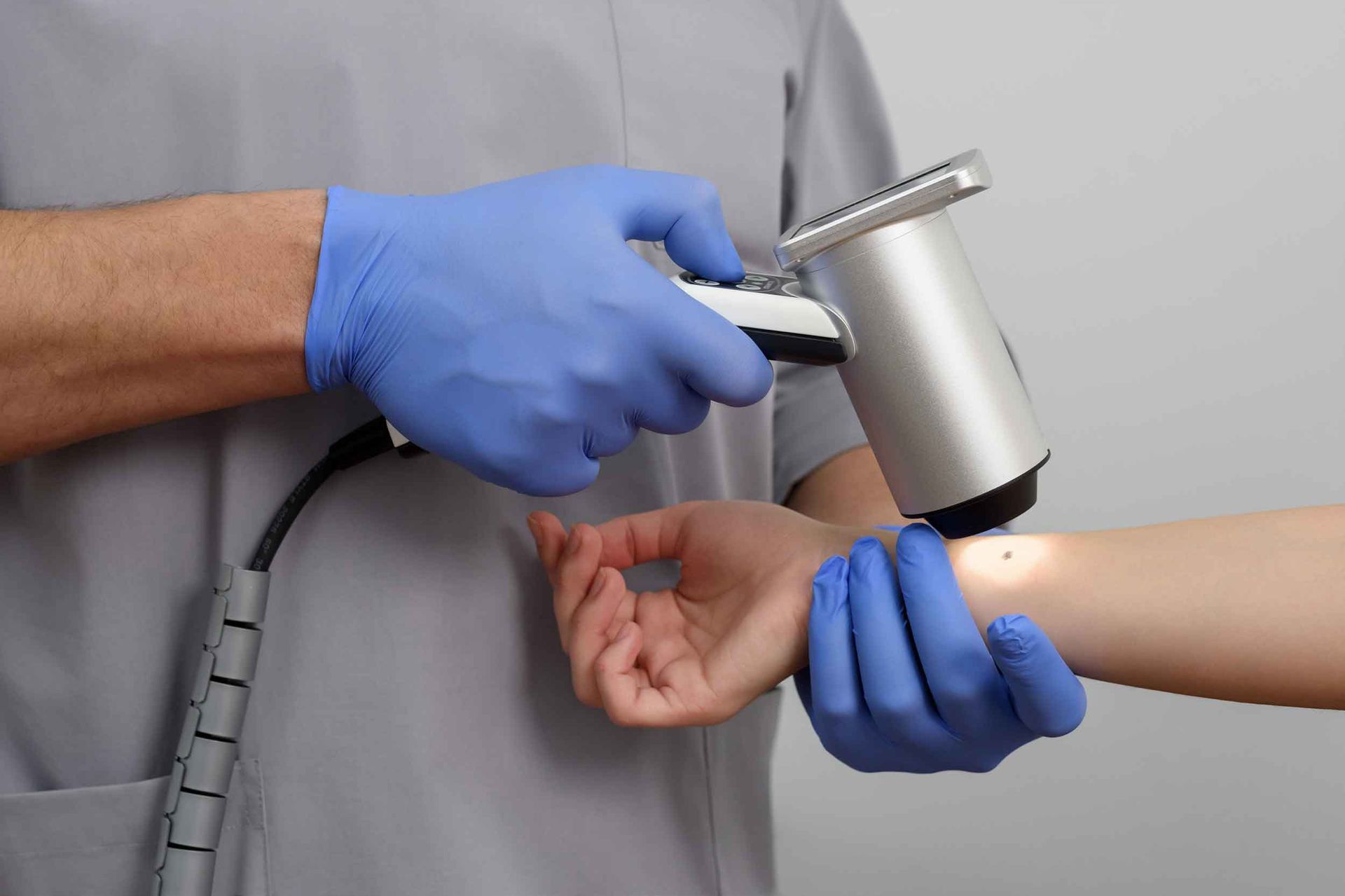 A skin specialist in blue gloves using a digital dermatoscope to examine a patient's mole on her arm.