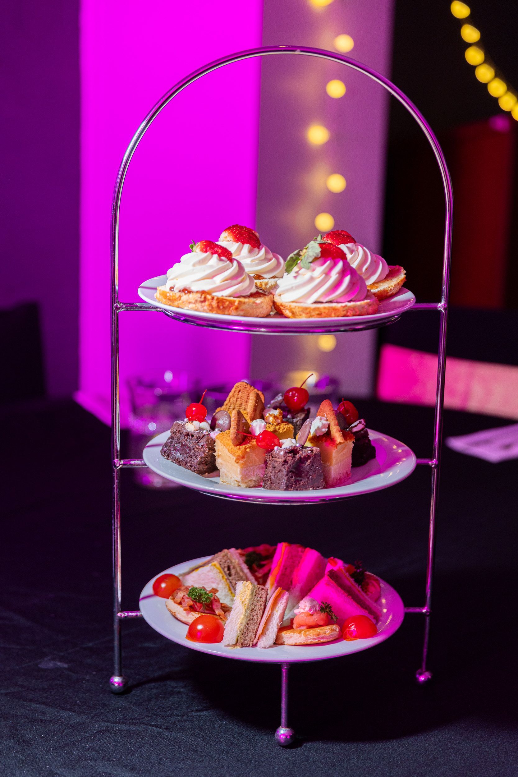 Three-tiered dessert stand with mini quiches, pastries, and sandwiches, lit by pink light.