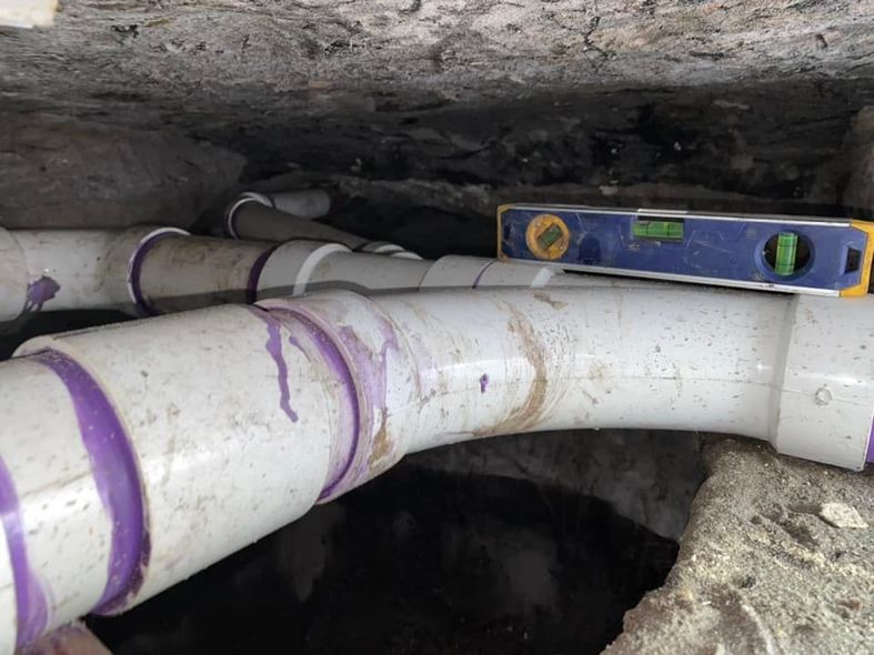 White PVC pipes with purple joints in a dark crawl space; a level is on one pipe.