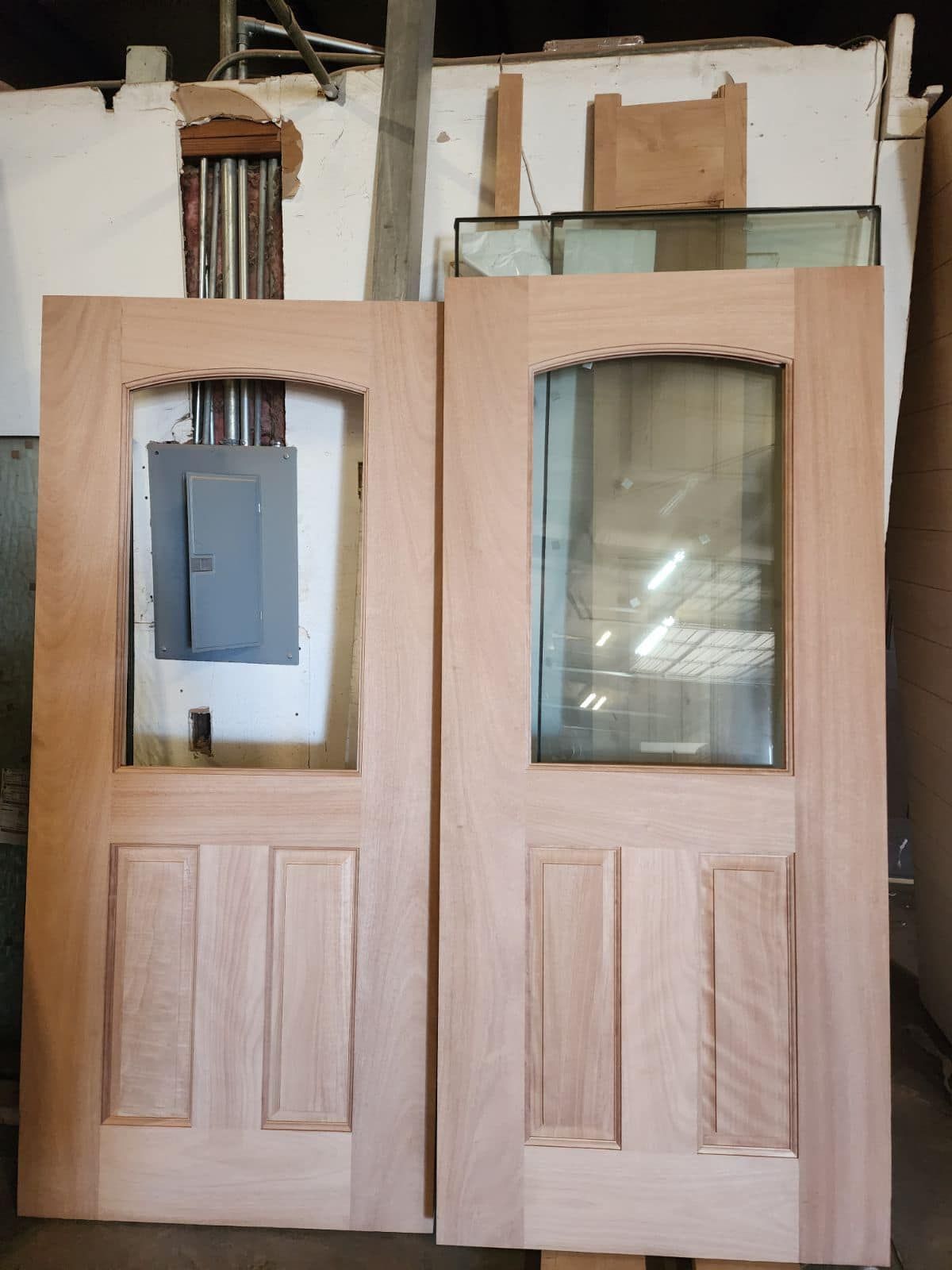 Two unfinished wooden doors with a glass pane, stacked against a wall with electrical box.