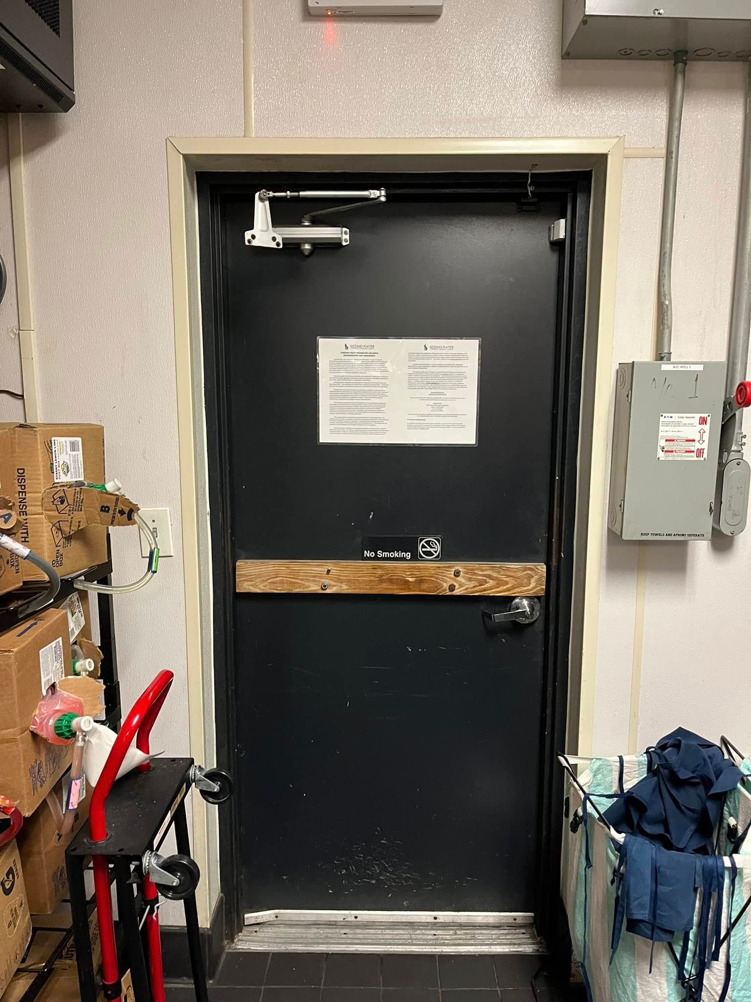 Black door with a sign, closer and bar, set in a utility room. Electrical box and supplies visible.