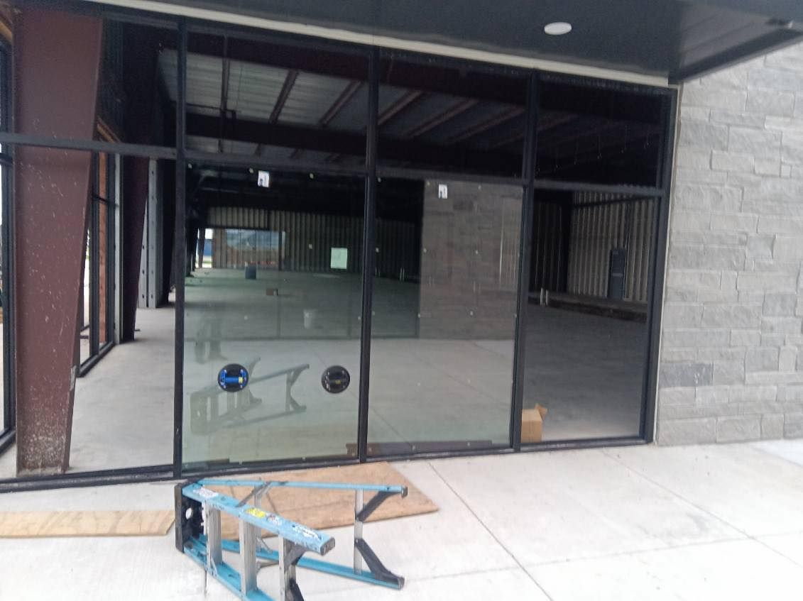Exterior view of an empty commercial space with large glass windows and concrete flooring; blue ladder present.