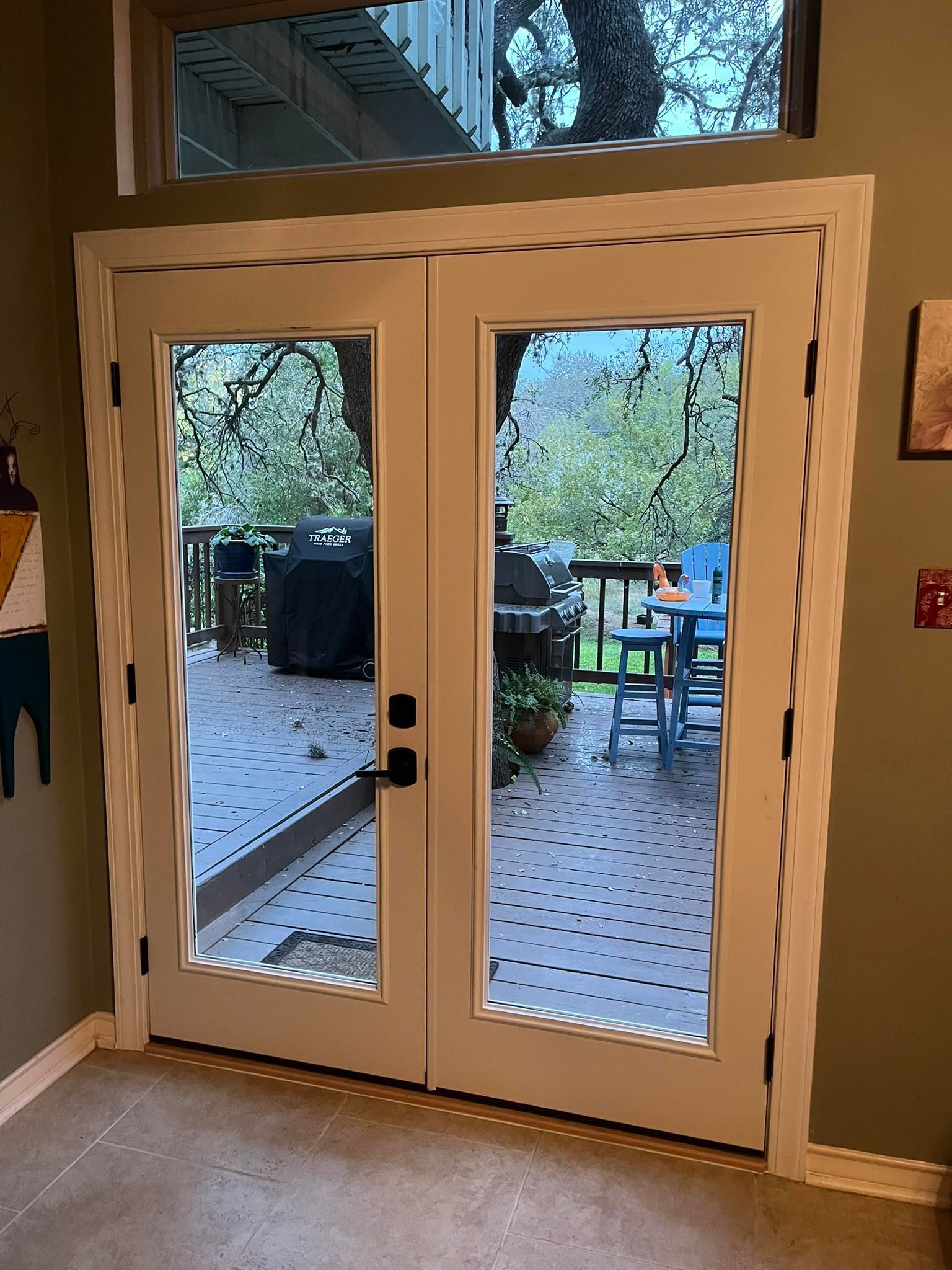 White French doors with glass panes, leading to a deck with outdoor furniture and a grill; a window above the doors.