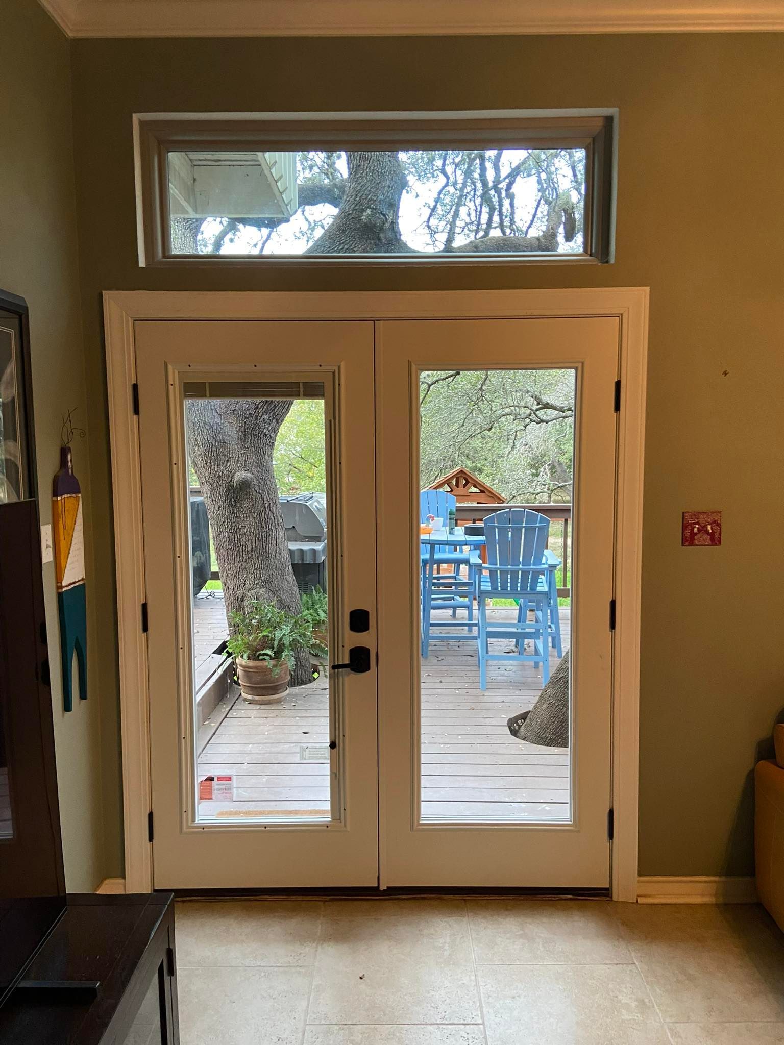 Double glass doors and transom window, opening to a deck with blue furniture and a tree; light interior walls.