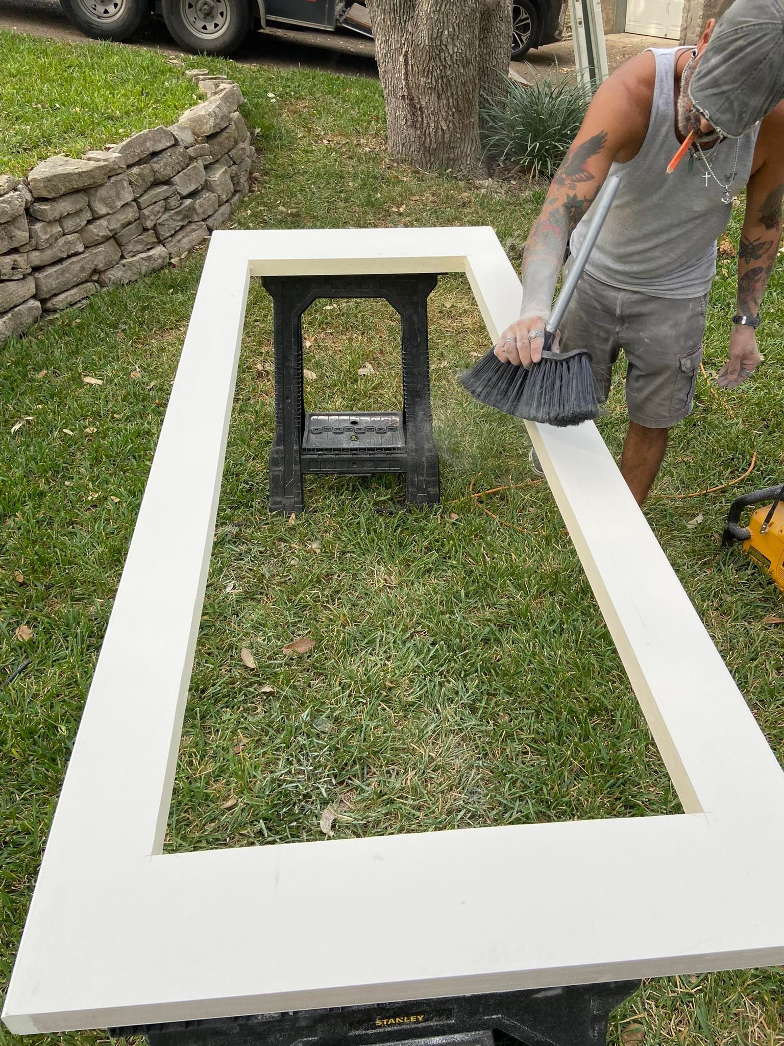 Person painting a white rectangular frame outdoors with a brush, grass and a sawhorse are in view.