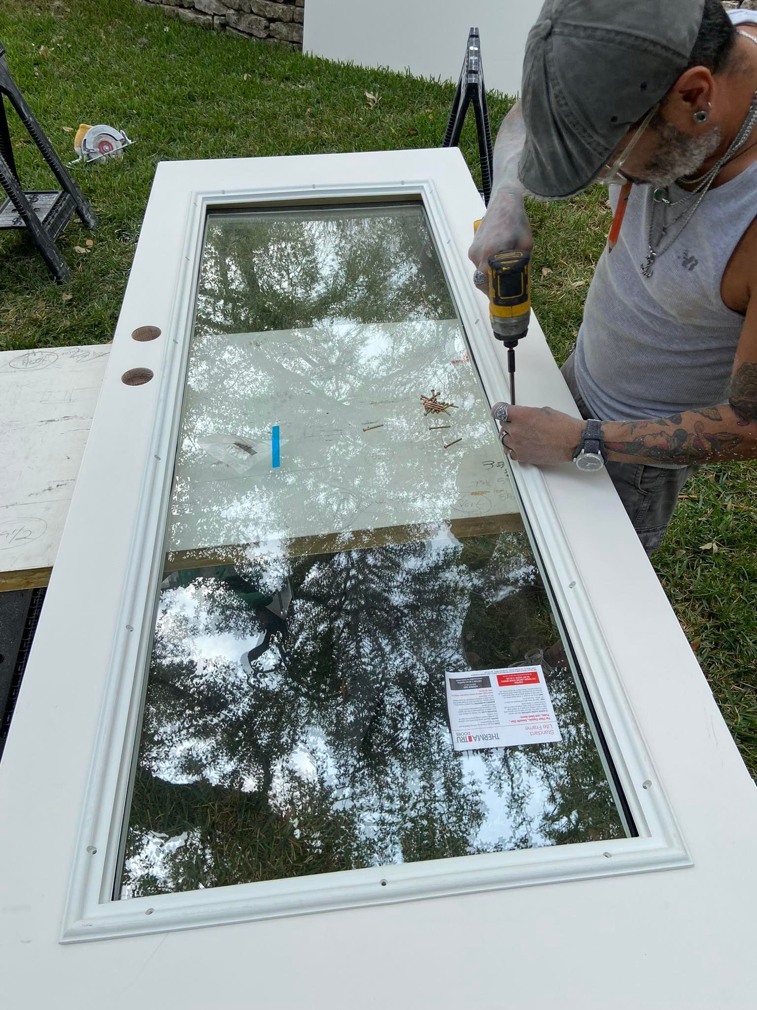 Person screws in window frame of white door outdoors, green grass in background.
